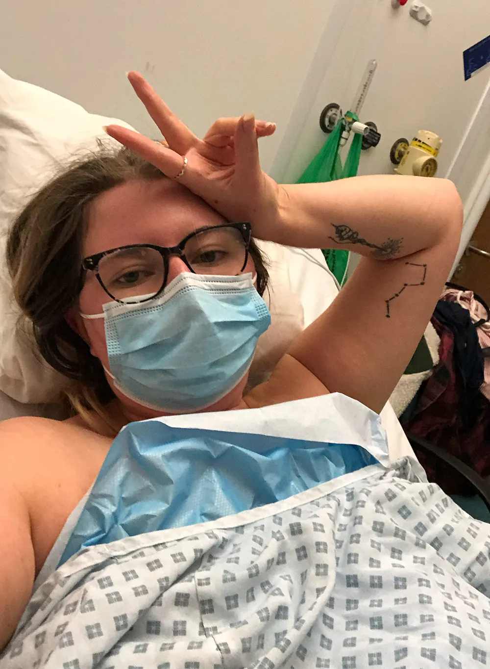Clare in a hospital bed flashing to peace sign.