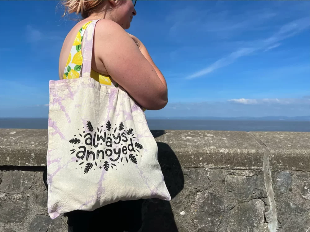 Woman holding an Always Annoyed Tote Bag on the seafront
